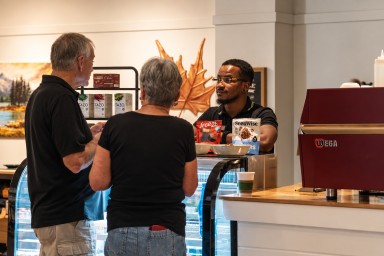 Two guests at the 2G2B Healthy caf&eacute; counter while a team member shows them free2b and SeenWise snacks